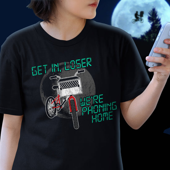 A model in a black tee shirt. On the front is a drawing of a red bike with a basket in the front. Above and below the bike is green text saying "Get in, loser, we're phoning home," stylized to look like text from old electronics. Behind the model is a full moon with a silhouette of a kid on a bike flying in front of it.