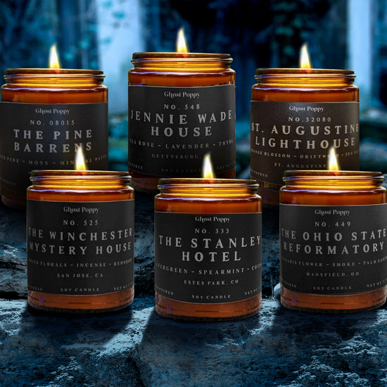 Six glass candles in two rows, sitting on a dark blue rock ledge in a cave. Each candle has a grey label with white text, listing the name of a famous haunted location.