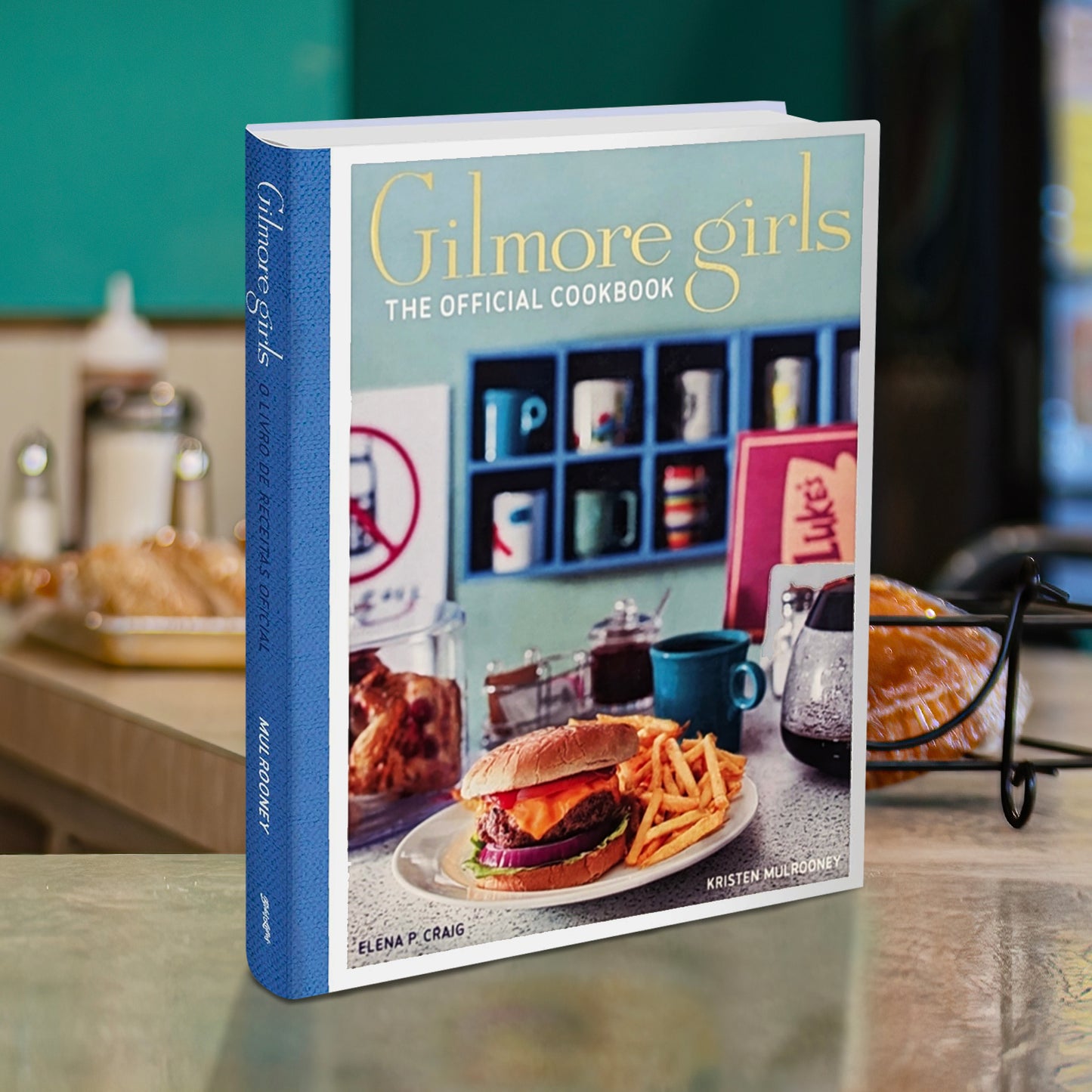 A mint green cookbook with a blue spine set against a vintage diner counter. The book cover reads "Gilmore Girls: The Official Cookbook" and features a plate of burgers and fries sitting in a vintage-style diner.