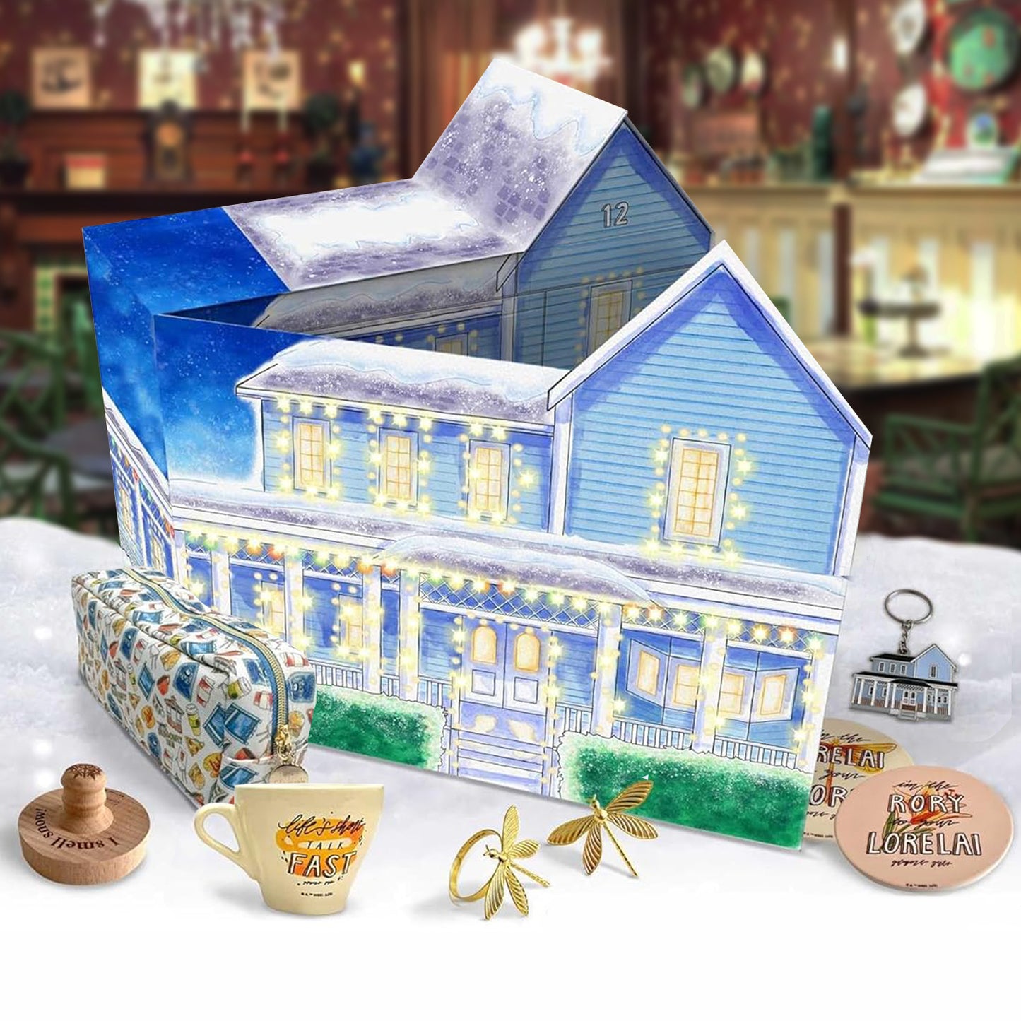 An image of a Gilmore Girls themed advent calendar sitting on a snowy bank. The calendar is shaped like a house, with various Gilmore Girls themed items in front of it. In the background is the Dragonfly Inn interior.