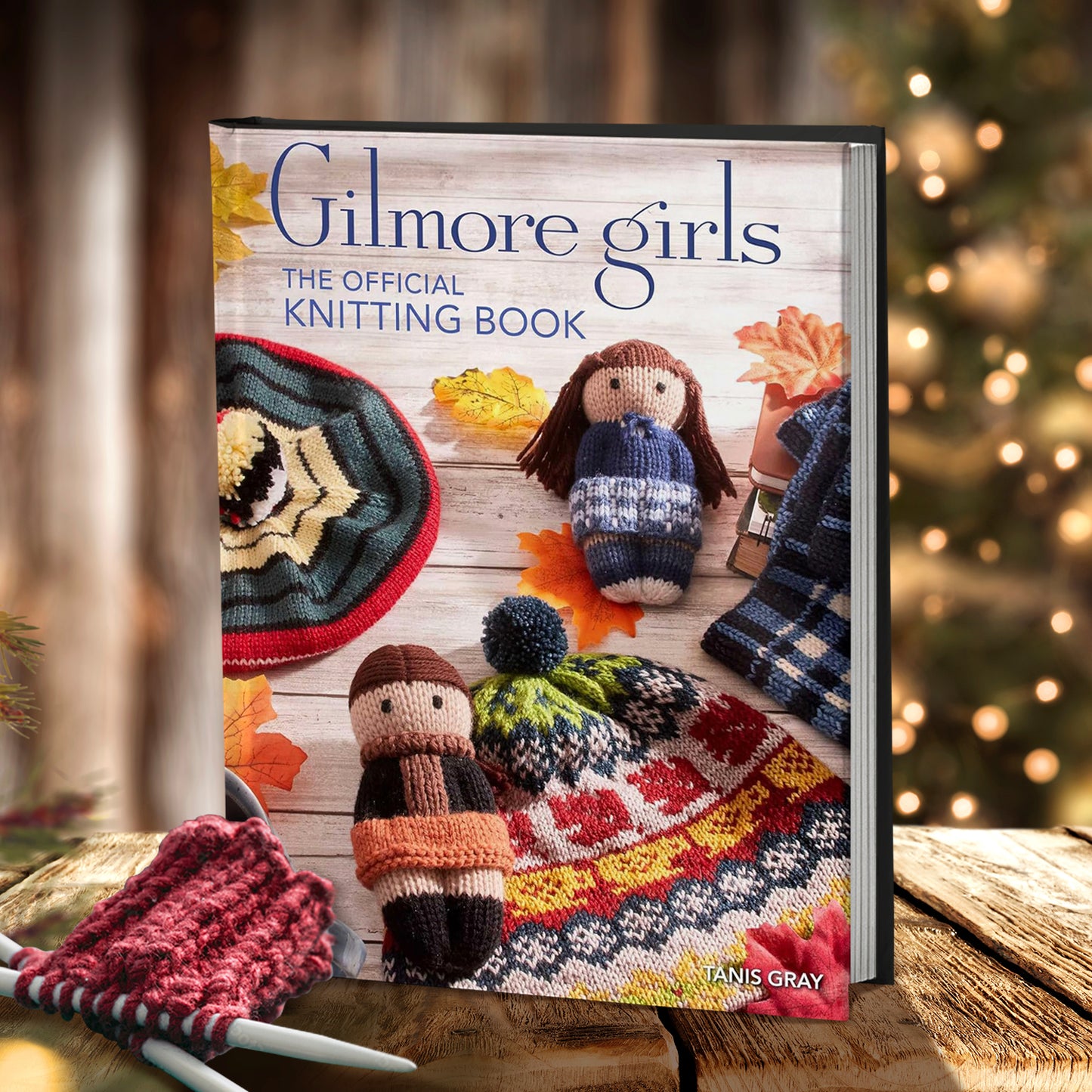 A hardcover book on a rough wooden table. The cover depicts knitted effigies of the main characters from the TV series The Gilmore Girls. Around them are other knitted items, including a hat with coffee mugs, a beret, and a blue plaid napkin. Blue text says "Gilmore Girls, the official knitting book." In front of the book are two white knitting needles and a swatch of maroon yarn. In the background is a blurred Christmas tree with white lights.