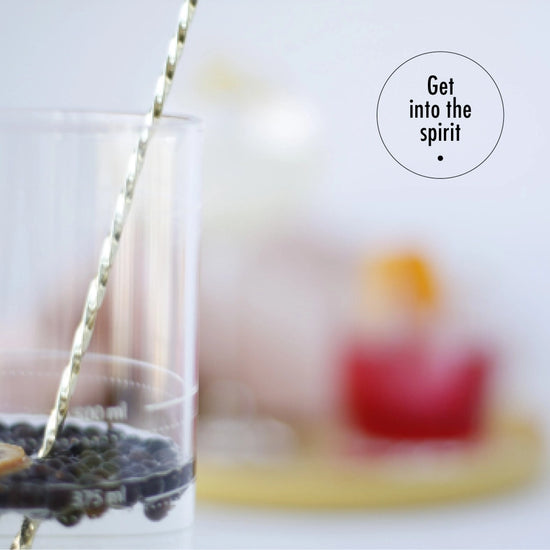 A highball cocktail glass with a metal stirring stick. Inside the glass is gin with a black spice floating at the top. In the background is a blurred cocktail glass with a red beverage in it. At the top right corner is a black circle with black text inside saying "Get into the spirit."