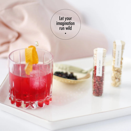 Load image into Gallery viewer, A cocltail glass with a red beverage and orange peel inside, standing on a white tray. Next to the glass are small vials filled with botanicals for cocktails. A pink pillow sits behind the tray. At the top is a black circle with black text inside saying &amp;quot;let your imagination run wild.&amp;quot;

