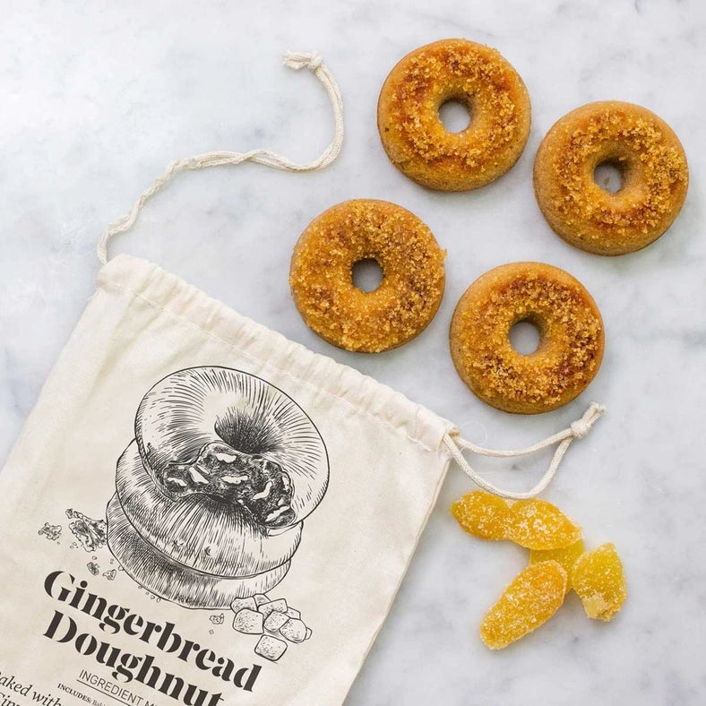Load image into Gallery viewer, A white canvas sack on a stone floor. On the sack is black text saying &amp;quot;Gingerbread Doughnut.&amp;quot; At the top is a black drawing of a stack of doughnuts. Next to the sack are four freshly-baked doughnuts and pieces of candied ginger.
