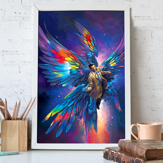 A framed painting against a white brick wall. The painting depicts the angel Castiel with rainbow wings, soaring through the nighttime sky. In front of the painting are old books, a wood jar of pencils, and a coffee mug.