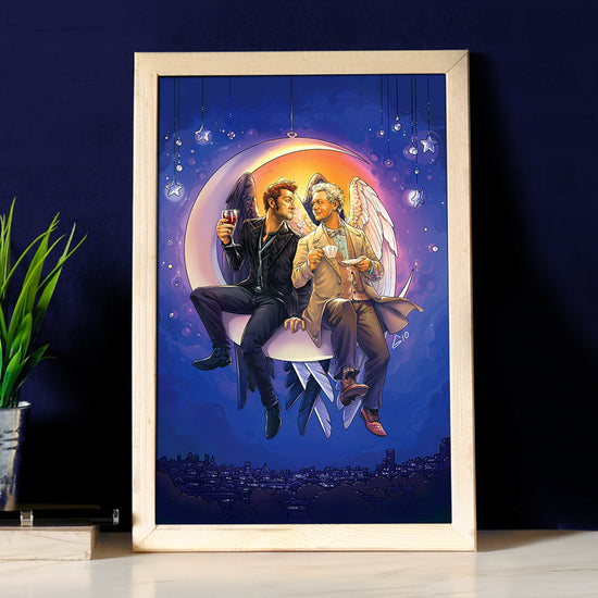 A framed painting on a wood table, against a dark blue background. The painting depicts Aziraphale and Crowley, from "Good Omens," smiling and sitting together on a crescent moon above a city skyline. Crowley holds a glass of wine, and Aziraphale holds a cup of tea. Next to the painting is a metal pot with a green plant