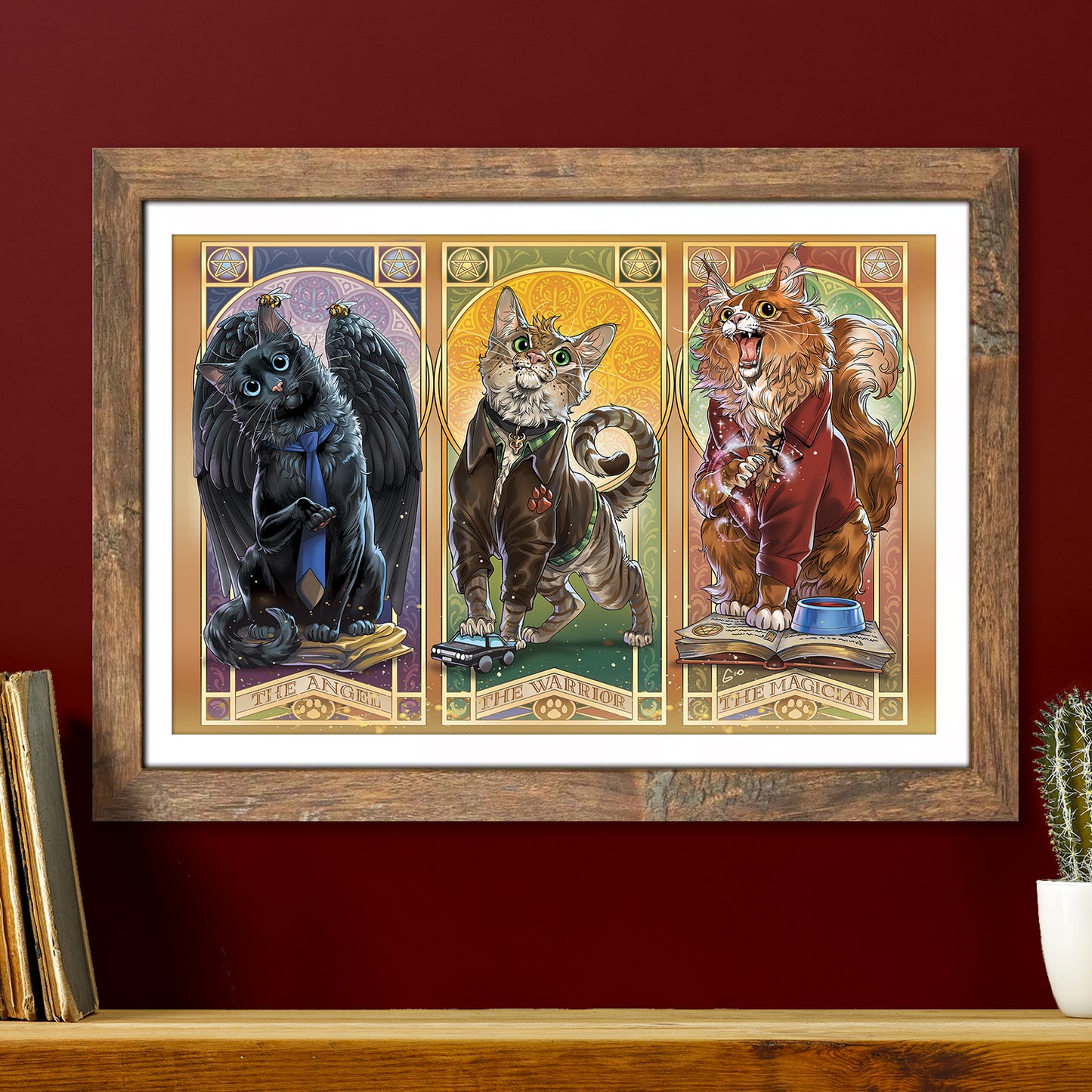 A framed lithograph against a maroon background. Under the frame is a bookshelf with a potted cactus and a two old books standing on it. The lithograph depicts three cats in separate panels. The first is a black cat with black wings and a blue necktie, named The Angel. The second is a tabby in a brown jacket, clutching a toy car, named The Warrior. The third is a Main Coon in a brown jacket, standing on an open book, named The Magician.