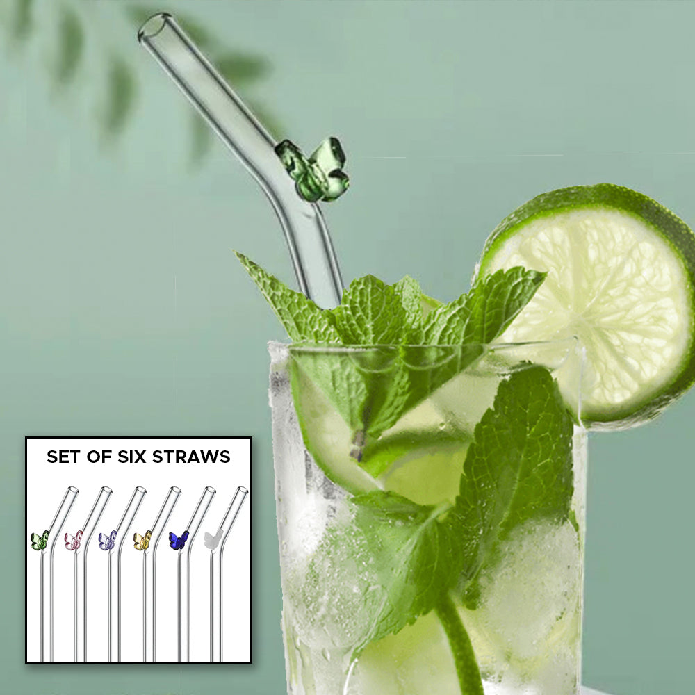 Close up of a tall glass filled with a clear beverage and lime slices. A glass drinking straw is sticking out of the top, with a small green glass butterfly attached at the straw's bend point. There is an inset image of six butterfly glass straws in green, pink, purple, yellow, blue, and white. Text in the image reads "set of six straws."