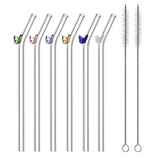 Six glass drinking straws against a white background. Each straw has a bend at the top, and a small glass butterfly is attached. Each butterfly is a different color. Next to the straws are two wire cleaning brushes with nylon bristles.