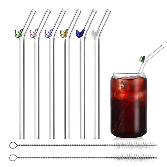 Five glass drinking straws against a white background. Each straw has a bend at the top, and a small glass butterfly is attached. Each butterfly is a different color. Below the straws are two wire cleaning brushes with nylon bristles. Next to the straws is a drinking glass filled with a dark beverage. A glass straw is sticking out of the top of the glass.