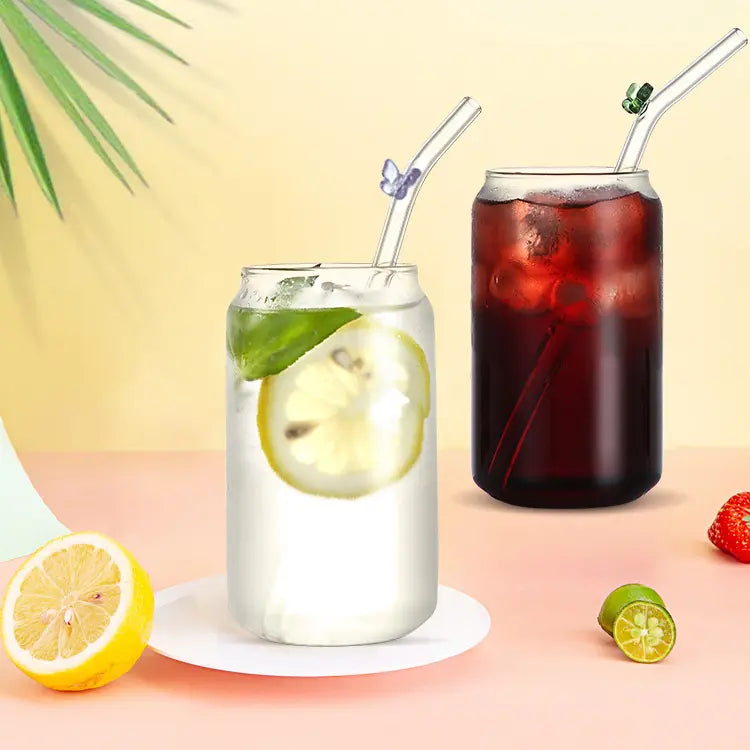 Two drinking glasses on a pink table. Each is filled with a different colored beverage. A glass drinking straw is sticking out of each glass. Each straw has a small colored butterfly attached to it.