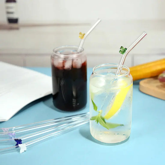 Two drinking glasses on a blue table. Each is filled with a different colored beverage. A glass drinking straw is sticking out of each glass. Each straw has a small colored butterfly attached to it. Four more glass straws are laying on the table between the two drinking glasses.