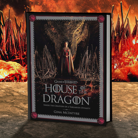 A black hardcover book on a stone table. The cover depicts a woman in a red and gold dress standing on a pile of swords. with a dragon's head directly behind her. Grey text says "Game of Thrones: House of the Dragon. inside the creation of a Targaryen dynasty." Around the edge is a grey border shaped like dragon teeth, with a red multi-headed dragon silhouette in each corner. Behind the book is the Iron Throne engulfed in flames.