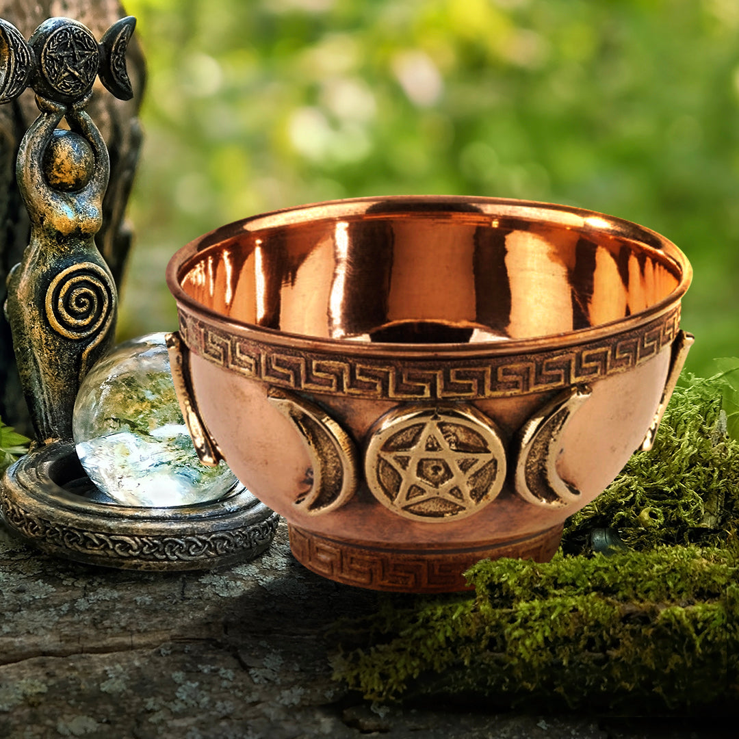 A copper bowl on a moss-covered log. The front of the bowl has half moons and a devil's trap symbol on it. Around the edges is a repeating geometric pattern. Next to the bowl is a brass statue with a glass sphere in the center. In the background is a blurred forest.