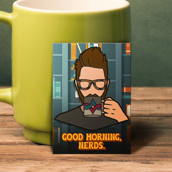 An enamel pin leaning against a green coffee mug. The pin has a stylized drawing of actor Wil Wheaton sipping from a coffee mug. The mug has a red and blue crossed line pattern against two shades of grey. At the bottom is yellow text saying "good morning, nerds."