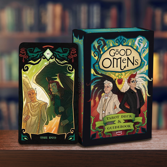 A black box on wood table, in front of bookshelves. At the top of the box is an oval with "good omens" in black text. Beneath the text is a drawing of Aziraphale and Crowley, the two main characters. Under them is a green banner with black text saying "tarot deck and guidebook." Behind the two characters, colored rays point upward and downward. A card from the deck stands next to the box, with a drawing of the two characters. Under them in green letters is "The duo."