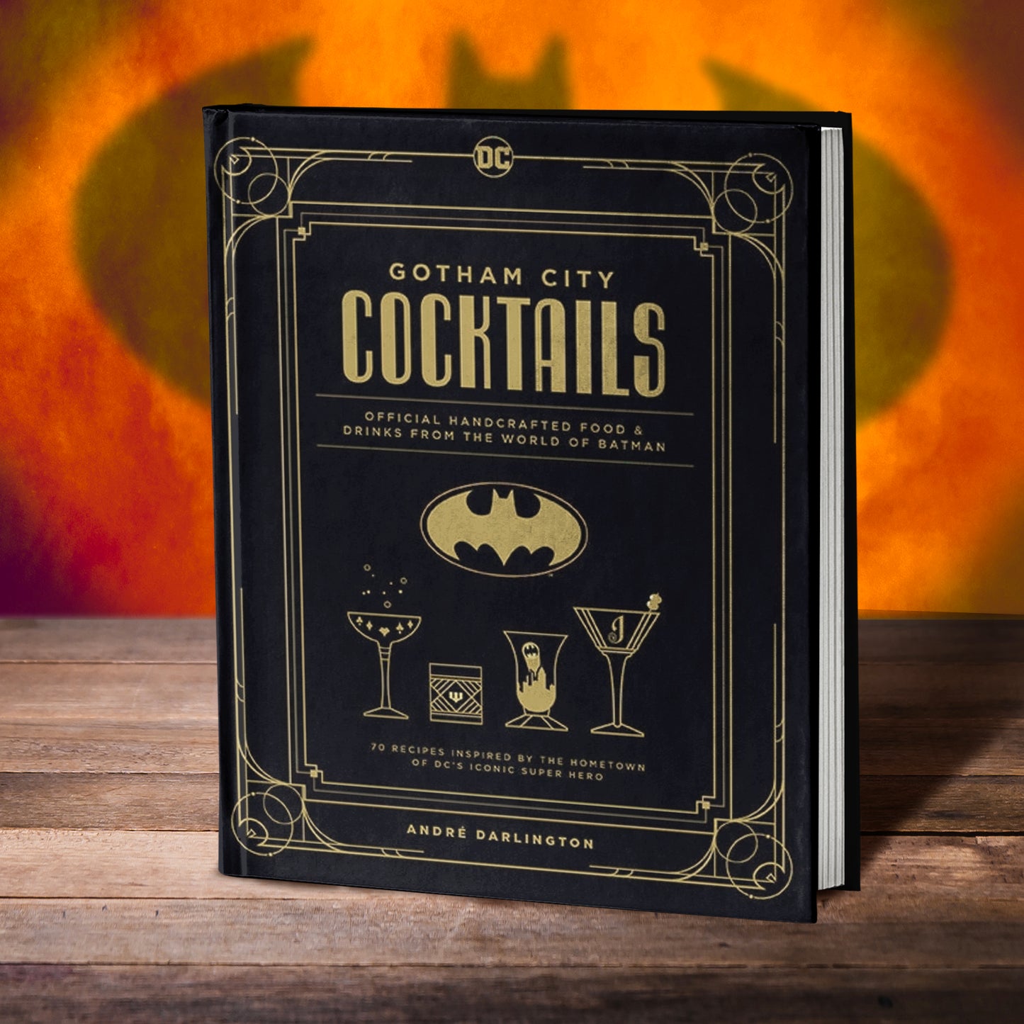 A black book on a wooden table.. Around the edges of the cover is an Art Deco border in yellow. Yellow text says "gotham city cocktails: official handcrafted food and drinks from the world of batman." Under the title is the Batman logo in yellow. At the bottom are line drawings of cocktail glasses. In the background is an orange and red wall, with a shadowy Batman symbol in the center.
