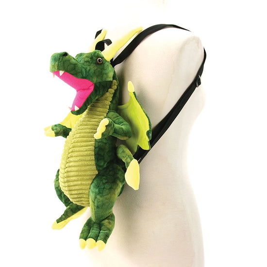 A dragon-shaped backpack in shades of green, hanging on a white mannequin. The dragon has green wings, and a red mouth, with black straps at the back.