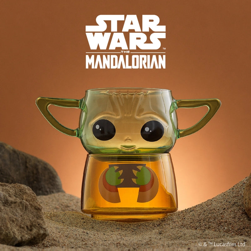 Two small drinking glasses stacked on each other on a stretch of sand, with an orange background. The two glasses combined depict the character Grogu from the Star Wars series The Mandalorian. The top glass has two handles shaped as his ears. At the top is white text saying "Start Wars, the Mandalorian."