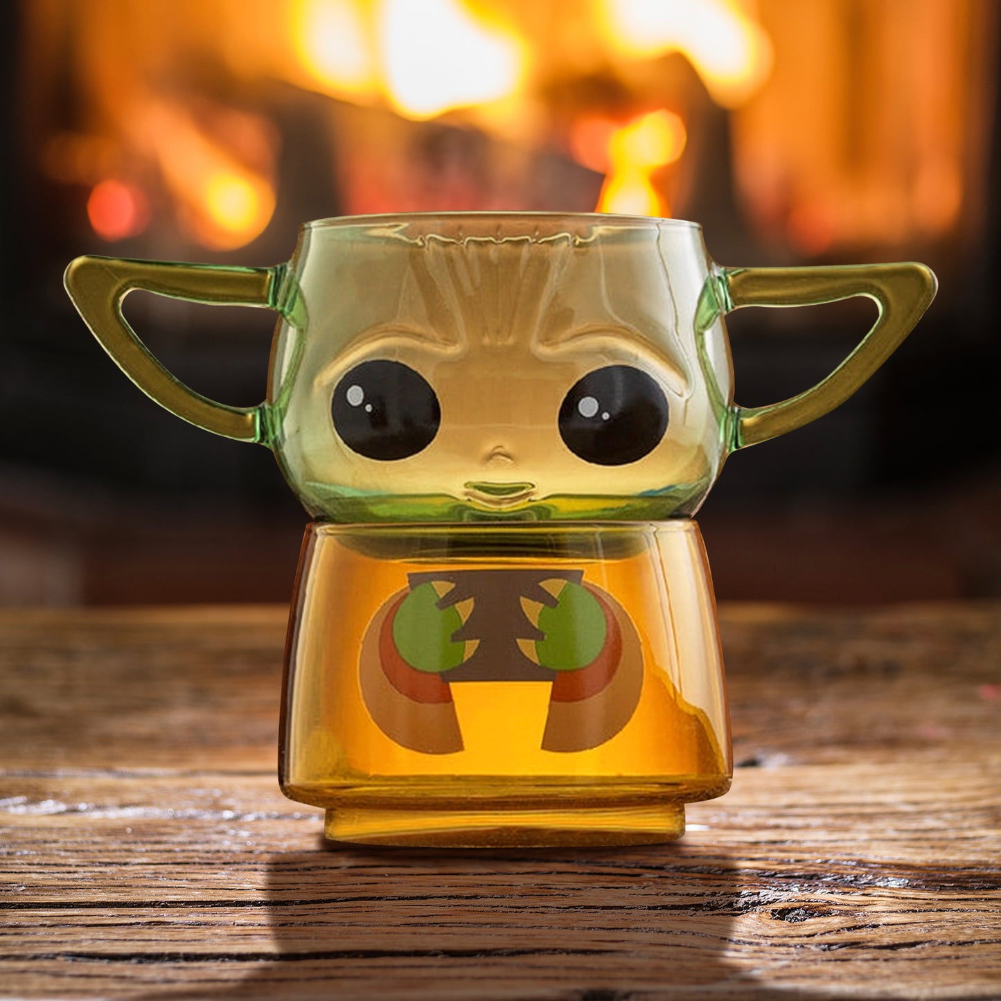 Two small drinking glasses stacked on each other on a wood table, with a fire in the background. The two glasses combined depict the character Grogu from the Star Wars series The Mandalorian. The top glass has two handles shaped as his ears.