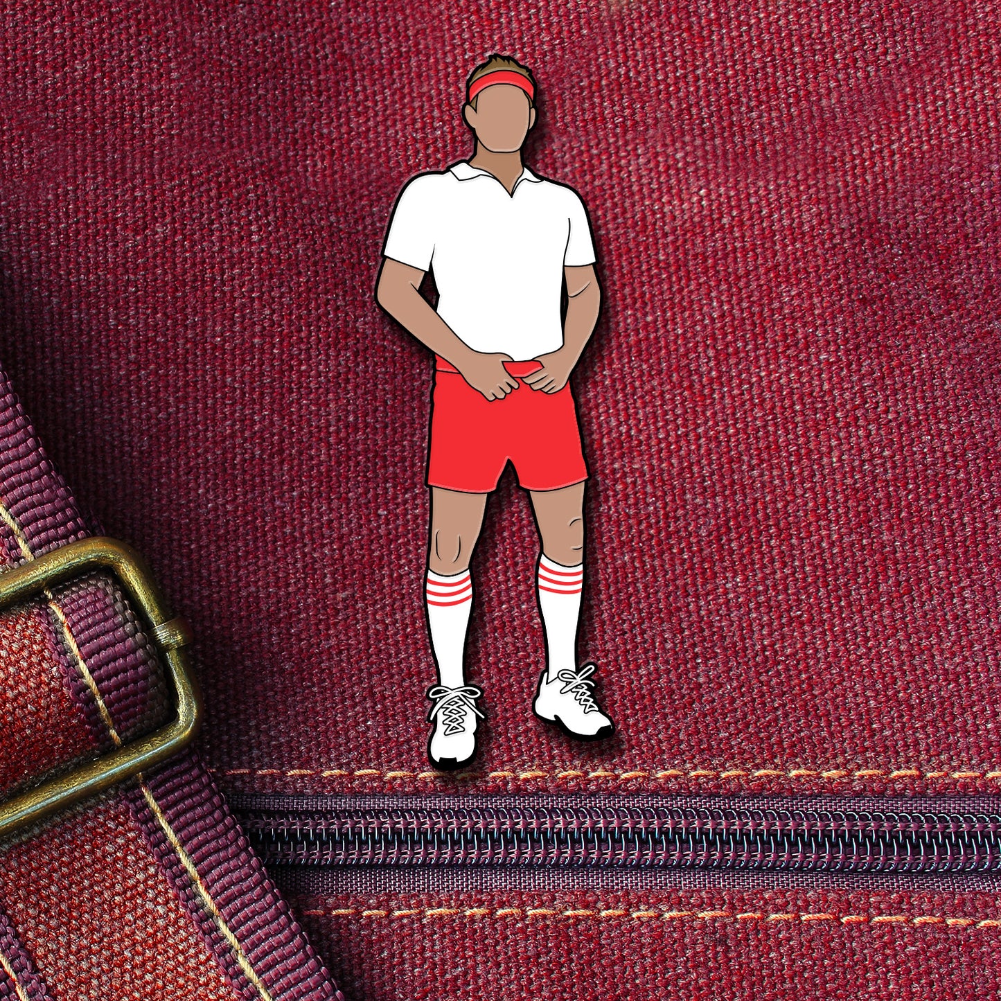 An enamel pin depicting Dean Winchester from the TV series "Supernatural." Sam is wearing red gym shorts and a white polo shirt, with athletic socks and a red headband. The pin is attached to a maroon canvas bag.