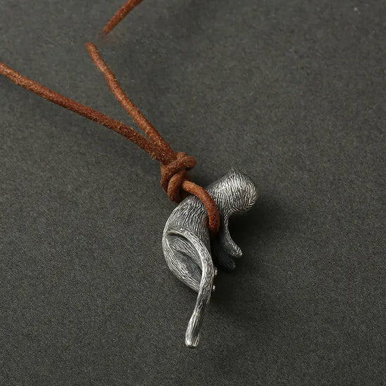 Close up of a cat-shaped pendant hanging from a brown leather cord, against a dark grey background. The cord is wrapped under the cat's abdomen, suspending it in mid-air.