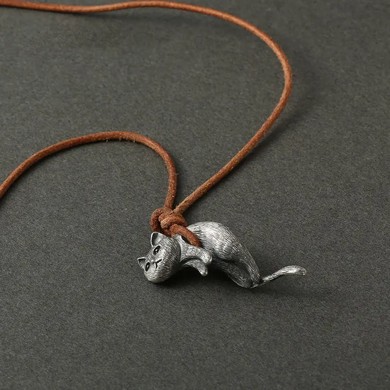 Close up of a cat-shaped pendant hanging from a brown leather cord, against a dark grey background. The cord is wrapped under the cat's abdomen, suspending it in mid-air.