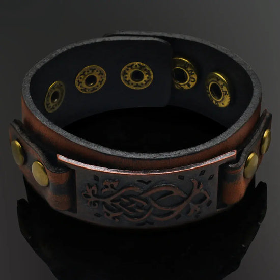 A dark brown leather cuff bracelet inspired by Viking imagery/style. It has brassy metal details, an adjustable button closure, and a brown metal plate with visual detailing.