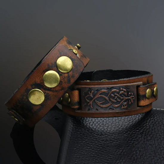 A dark brown leather cuff bracelet inspired by Viking imagery/style. It has brassy metal details, an adjustable button closure, and a brown metal plate with visual detailing.