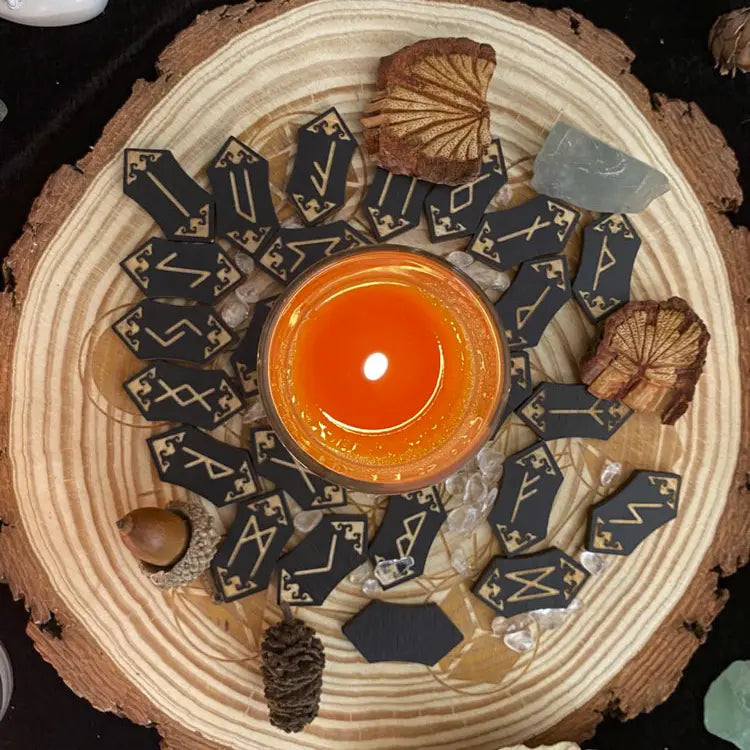 Load image into Gallery viewer, 24 cards, pointed on both ends, scattered around a flat tree stump. Each card is black, with a Nordic rune printed on it in gold. An orange candle burns in the center of the cards.
