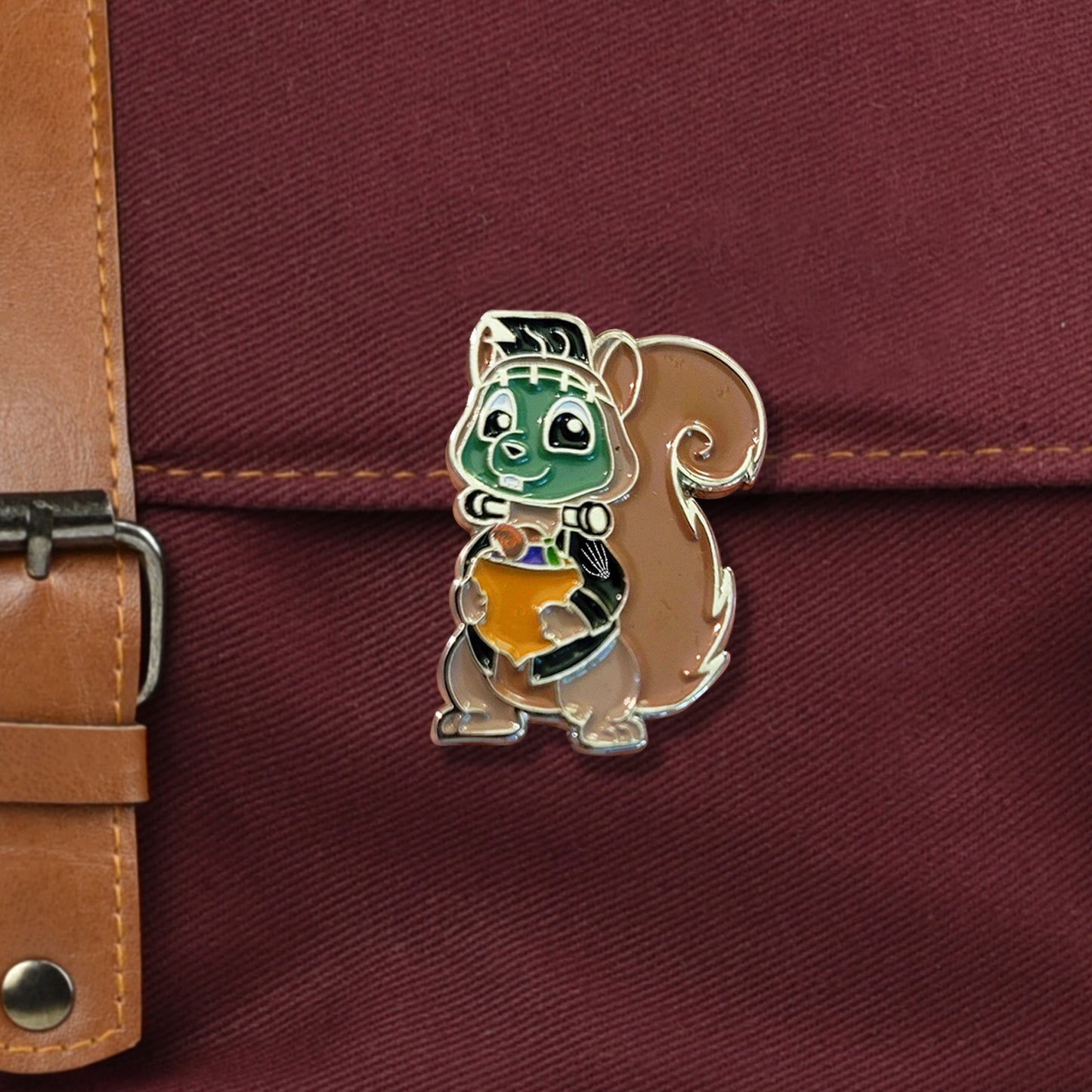 A closeup on a red bag with metal buckle. There is a medium-sized pin of a squirrel with green Frankenstein face paint and neck bolts holding an acorn full of candy. There is a small skeleton handprint on his left shoulder. 