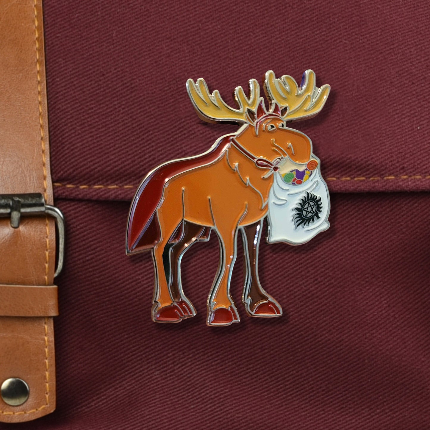 A closeup on a red bag with metal buckle. There is a large pin of a moose in a devil costume carrying a sack full of candy with an anti-possession symbol on it.