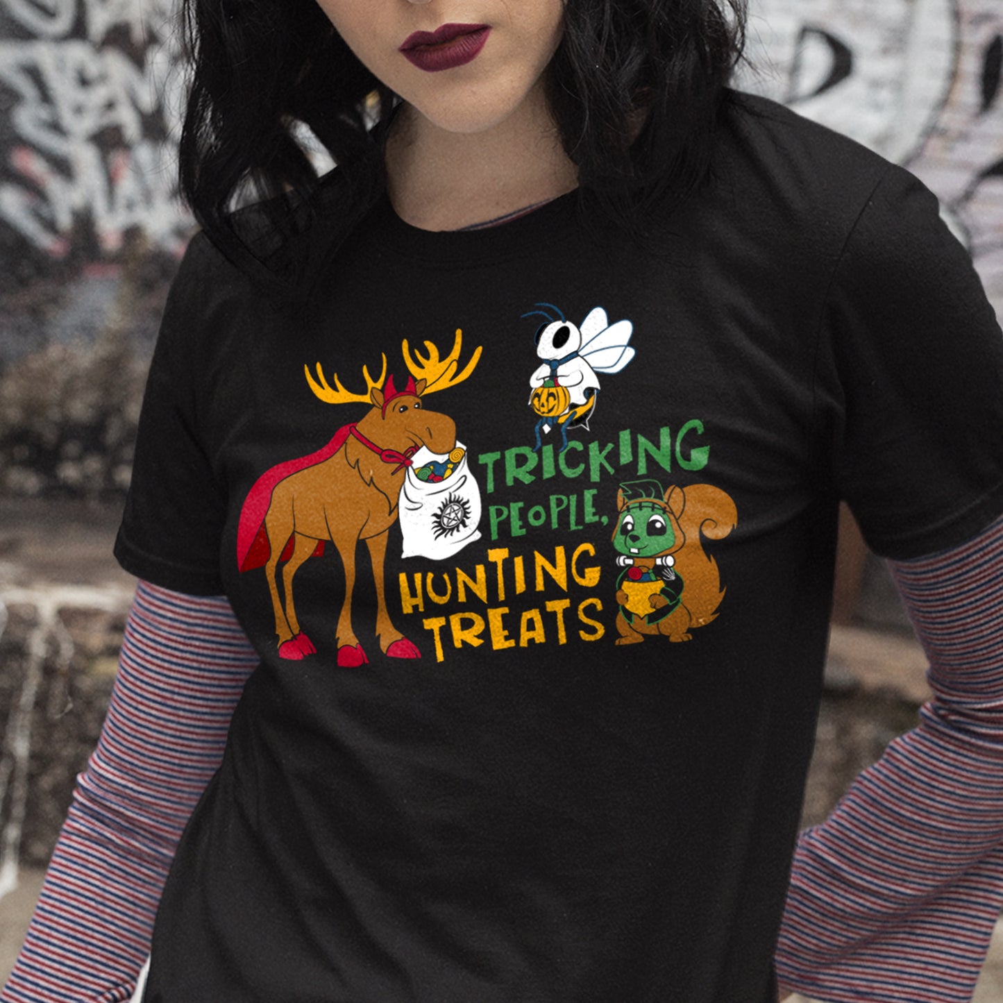 A female model wearing a black t-shirt. On the front are a three cartoon characters, a large moose in a devil costume, a squirrel in a Frankenstein costume, and a bee in a ghost costume. The moose has a bag in its mouth, the squirrel has a an acorn, and the bee has a jack-o-lantern, all filled with candy. Yellow and green text says "tricking people, hunting things."