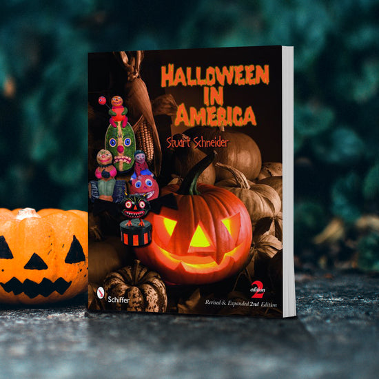 A dark book, with "Halloween in America" at the top in orange text. A brightly lit jack-o-lantern sits in the middle of the cover, currounded by brown pumpkins. On the left are various halloween-themed items. The book is standing on a black stone floor, with a jack-o-lantern next to it, and darkly lit green leaves in the background.