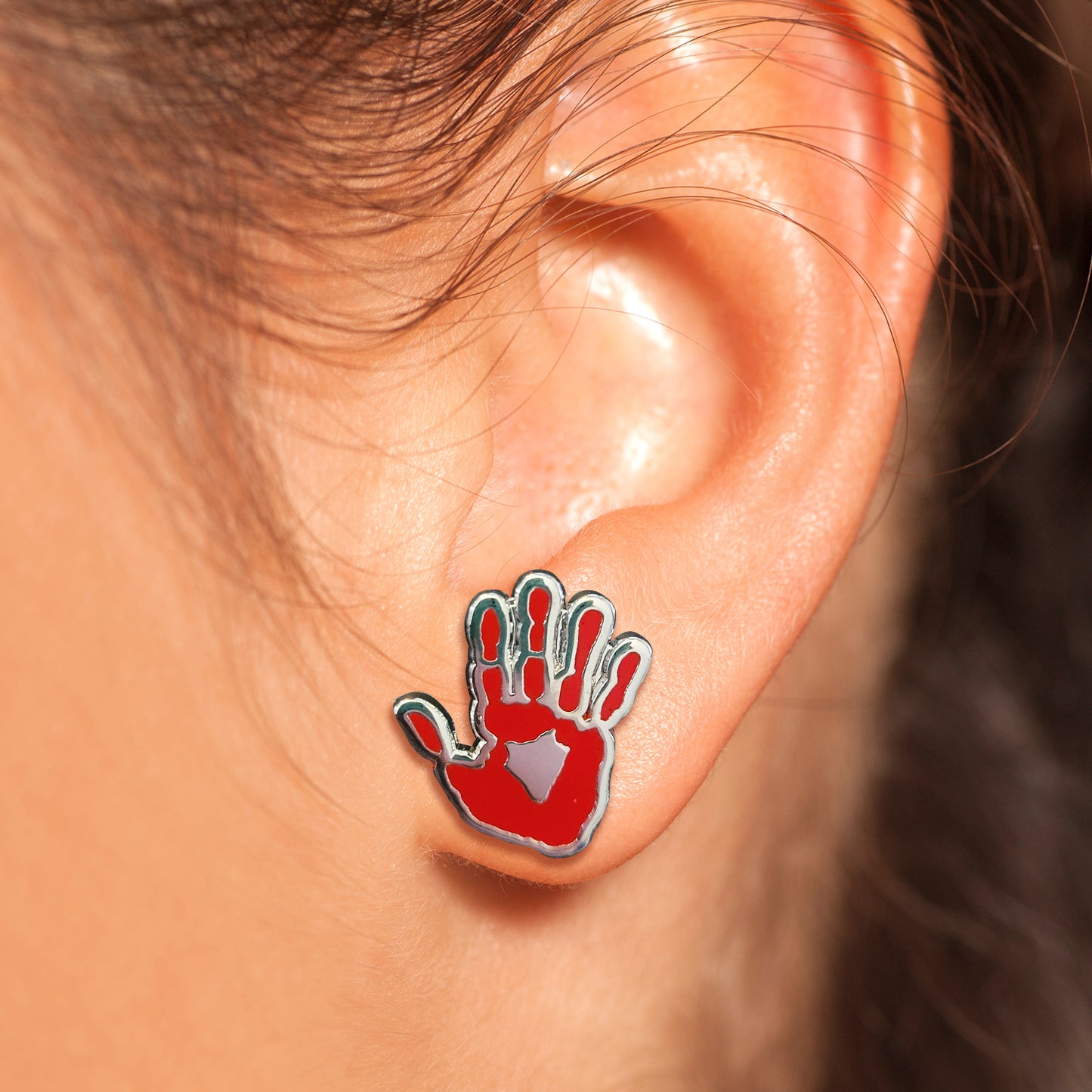 Close up of a model's ear, wearing an earring. The earring is shaped like a hand, painted red.