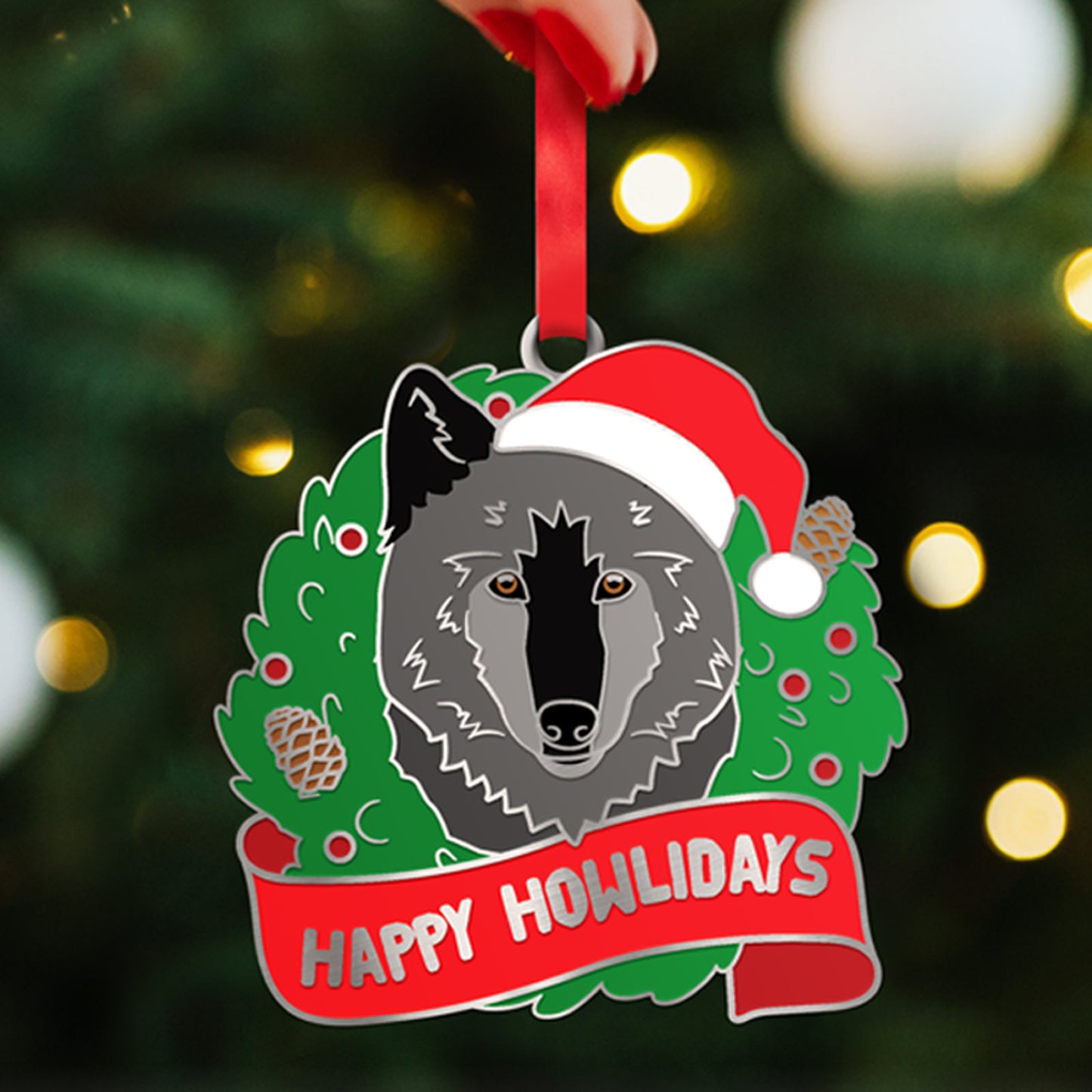 Close up of a Christmas ornament in front of a lit Christmas tree. The ornament depicts a grey wolf inside a green Christmas wreath. On the wolf's head is a red and white Santa hat. At the bottom is a red banner with silver text saying "Happy Howlidays."