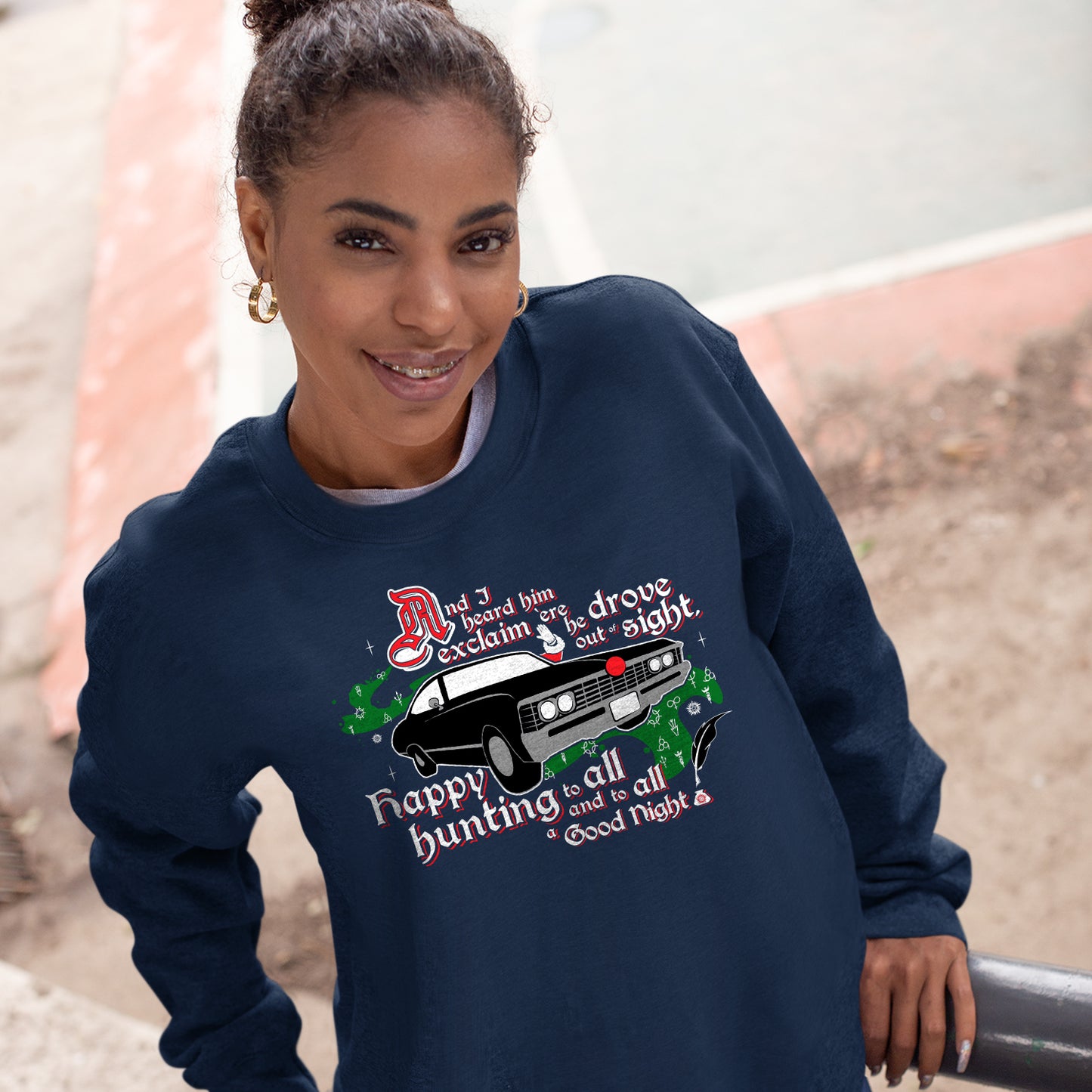 A woman wearing a navy blue crewneck sweatshirt. The sweatshirt reads "And I heard him exclaim 'ere he drove out of sight, happy hunting to all and to all a good night". In the middle of the words is a 3/4 view of a vintage Impala "wearing" a red nose in the front grill.  