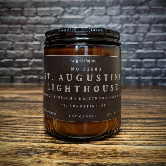 A glass candle jar on a wooden surface, with a brick wall in the background. The candle jar has a black label, with grey text saying "St. Augustine lighthouse. orange blossom, driftwood, sea salt. St. Augustine, FL."