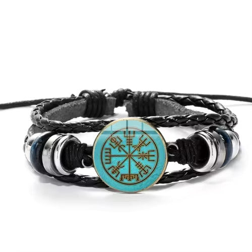 Close up of a black braided bracelet on a white background. At the center is a blue and gold round charm depicting Nordic runes in the shape of a compass.