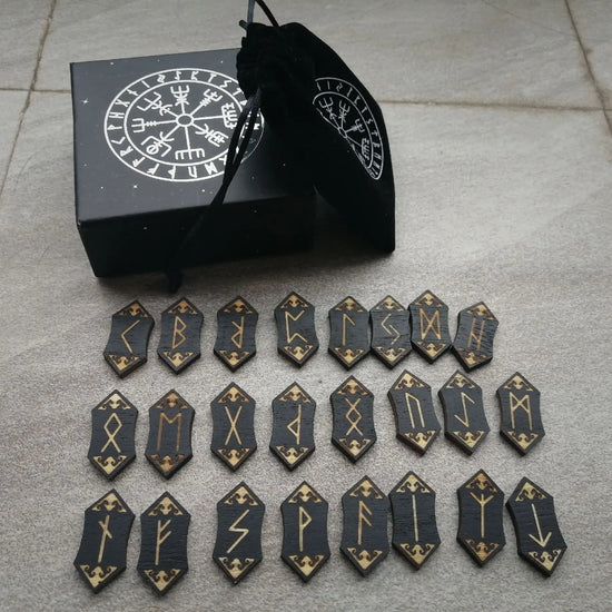 24 cards, pointed on both ends, in rows on a marble floor. Each card is black, with a Nordic rune printed on it in gold. Next to the cards is a black box with the Runic alphabet printed on it in silver, arranged in a circle. A black felt bag is leaning against the box.