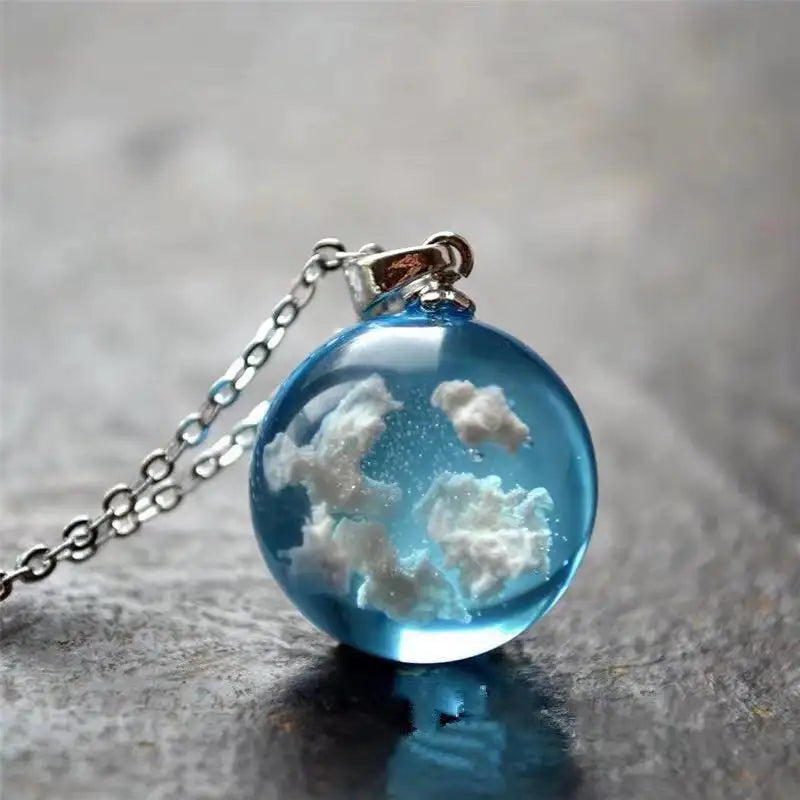 A round, sky-blue pendant on a dark stone table. The pendant is a sphere, with white clouds inside it. A silver chain is attached at the top.