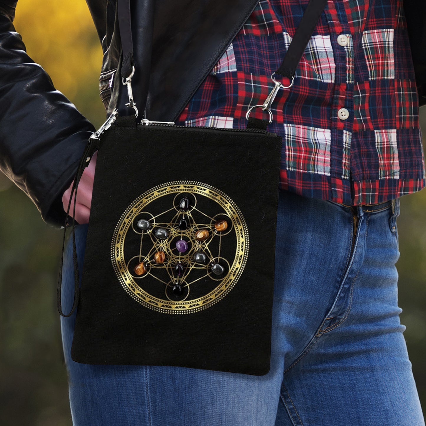 A small black crossbody bag on a female model. On the front of the bag is the Heaven's Portal symbol in gold ink, with gemstones inset in the circles. The bag has a small wrist strap attached at one side, and a larger shoulder strap at the top.