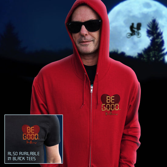 An image of actor Henry Thomas in a red zip hoodie. On the top left of the shirt is a red heart. On top of the heart is "Be Good" in yellow text, stylized to look like text from old electronics. Behind the model is a full moon with a silhouette of a kid on a bike flying in front of it. There is a small inset image of a man's chest wearing a black shirt with the same "Be Good" design on it. The inset reads "Also available in black tees."