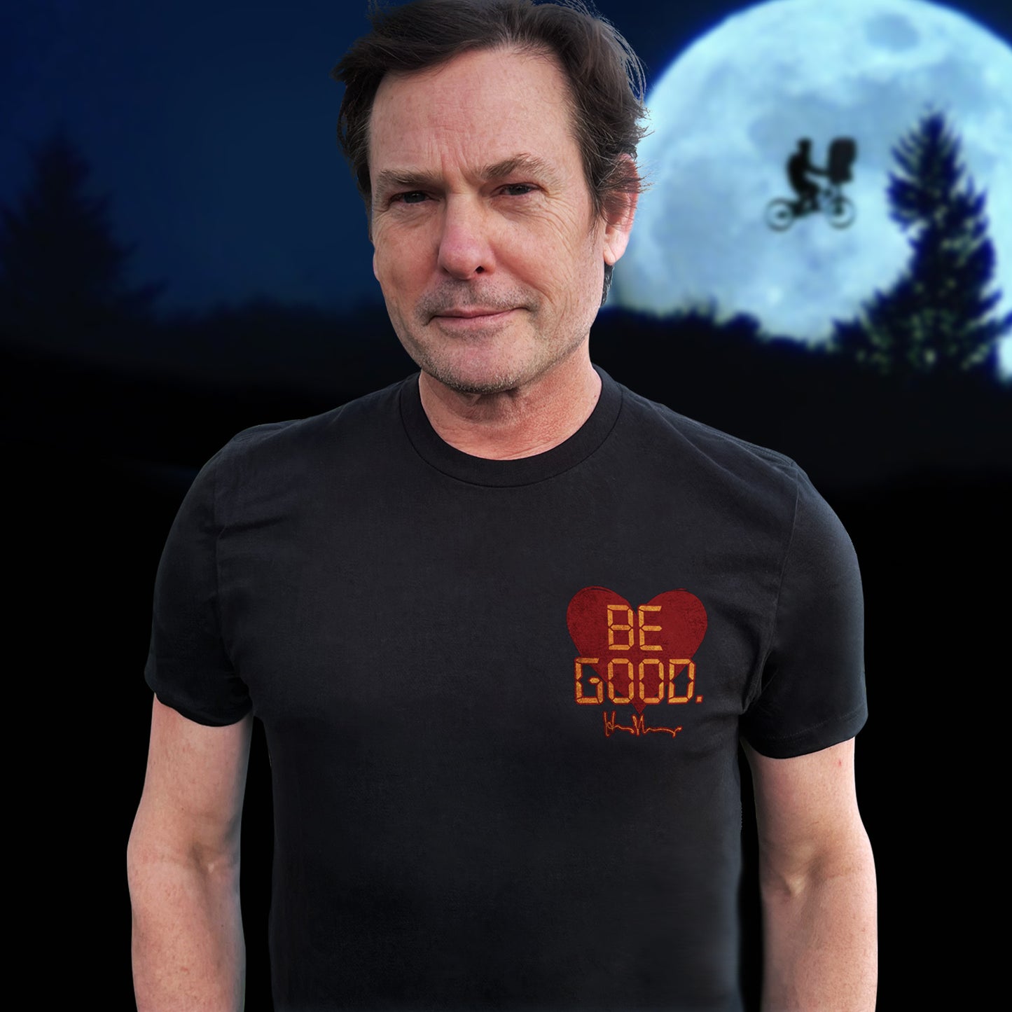 An image of actor Henry Thomas in a black tee shirt. On the top left of the shirt is a red heart. On top of the heart is "Be Good" in yellow text, stylized to look like text from old electronics. Behind the model is a full moon with a silhouette of a kid on a bike flying in front of it.