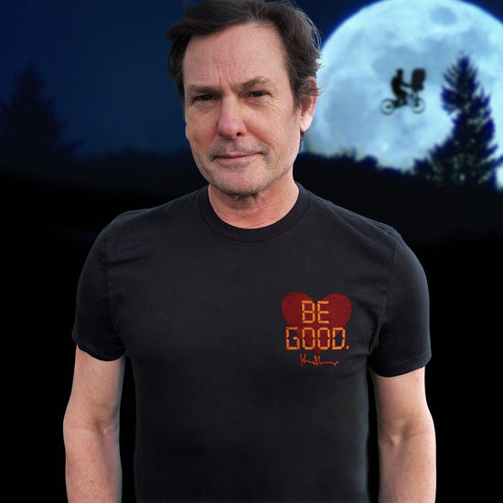 An image of actor Henry Thomas in a black tee shirt. On the top left of the shirt is a red heart. On top of the heart is "Be Good" in yellow text, stylized to look like text from old electronics. Behind the model is a full moon with a silhouette of a kid on a bike flying in front of it.