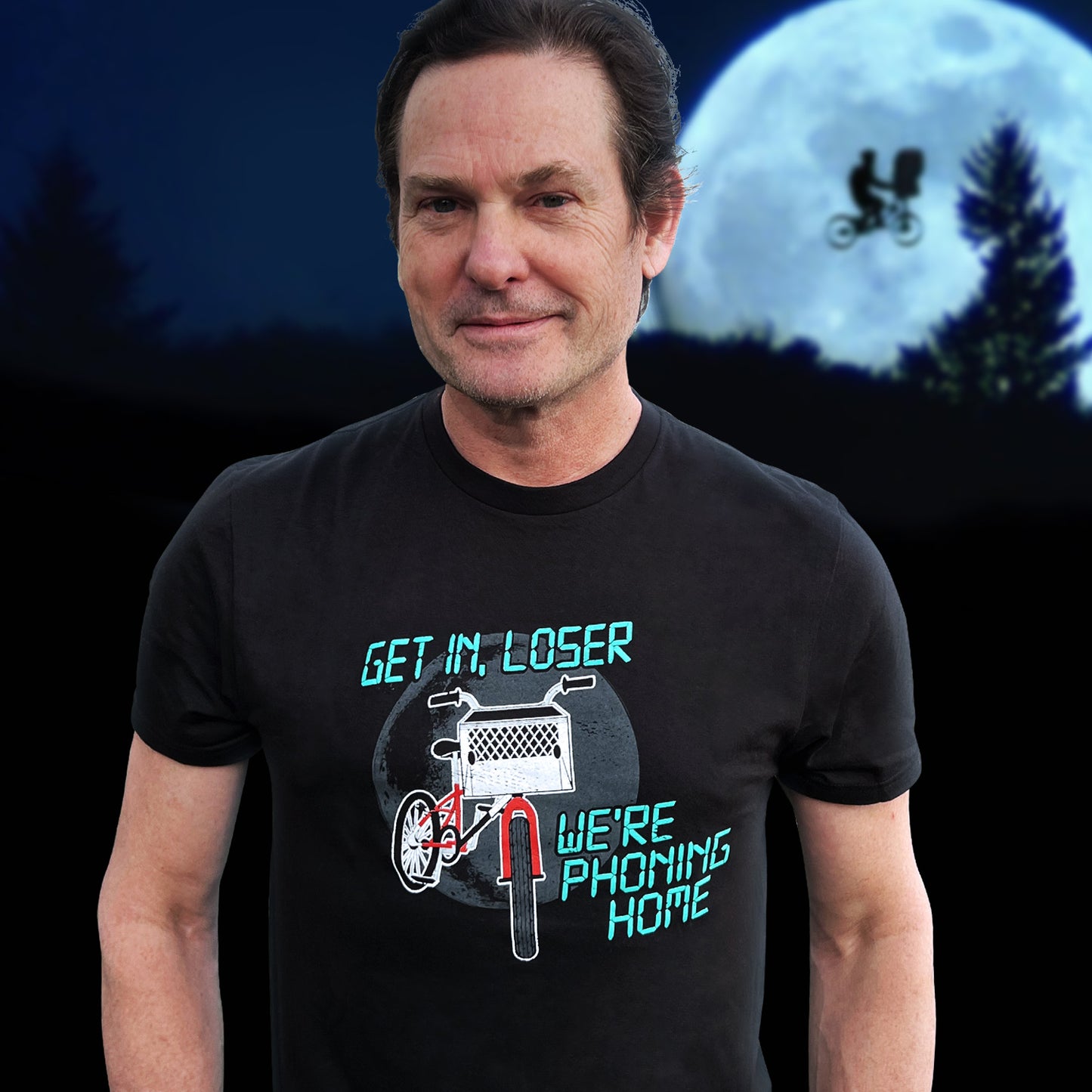 An image of actor Henry Thomas in a black tee shirt. On the front is a drawing of a red bike with a basket in the front. Above and below the bike is green text saying "Get in, loser, we're phoning home," stylized to look like text from old electronics. Behind Henry is a full moon with a silhouette of a kid on a bike flying in front of it.