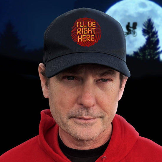 An image of actor Henry Thomas wearing a black cap with 'I'll be right here' on the front, with a red fingerprint under it. In the background is the night sky with moon and trees.