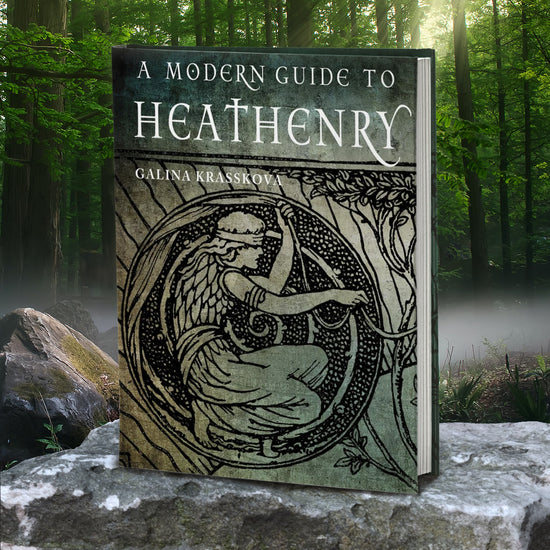 A grey book cover propped on a flat rock, against a forest background. At the top in white text is "A Modern Guide to Heathenry by Galina Krasskova." On the cover is a black line drawing of a pagan wearing a blindfold, crouching within a circle made from tree roots.
