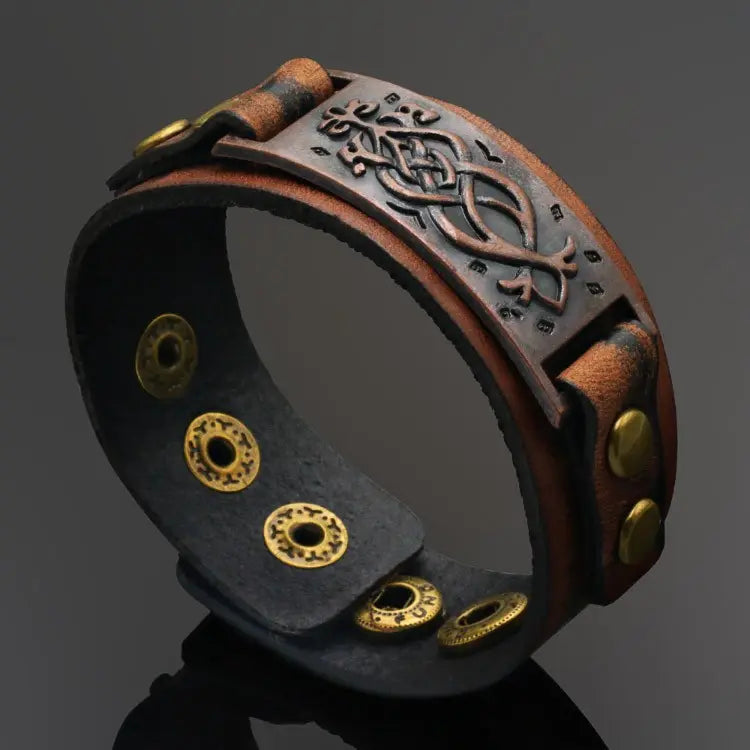 A dark brown leather cuff bracelet inspired by Viking imagery/style. It has brassy metal details, an adjustable button closure, and a brown metal plate with visual detailing.