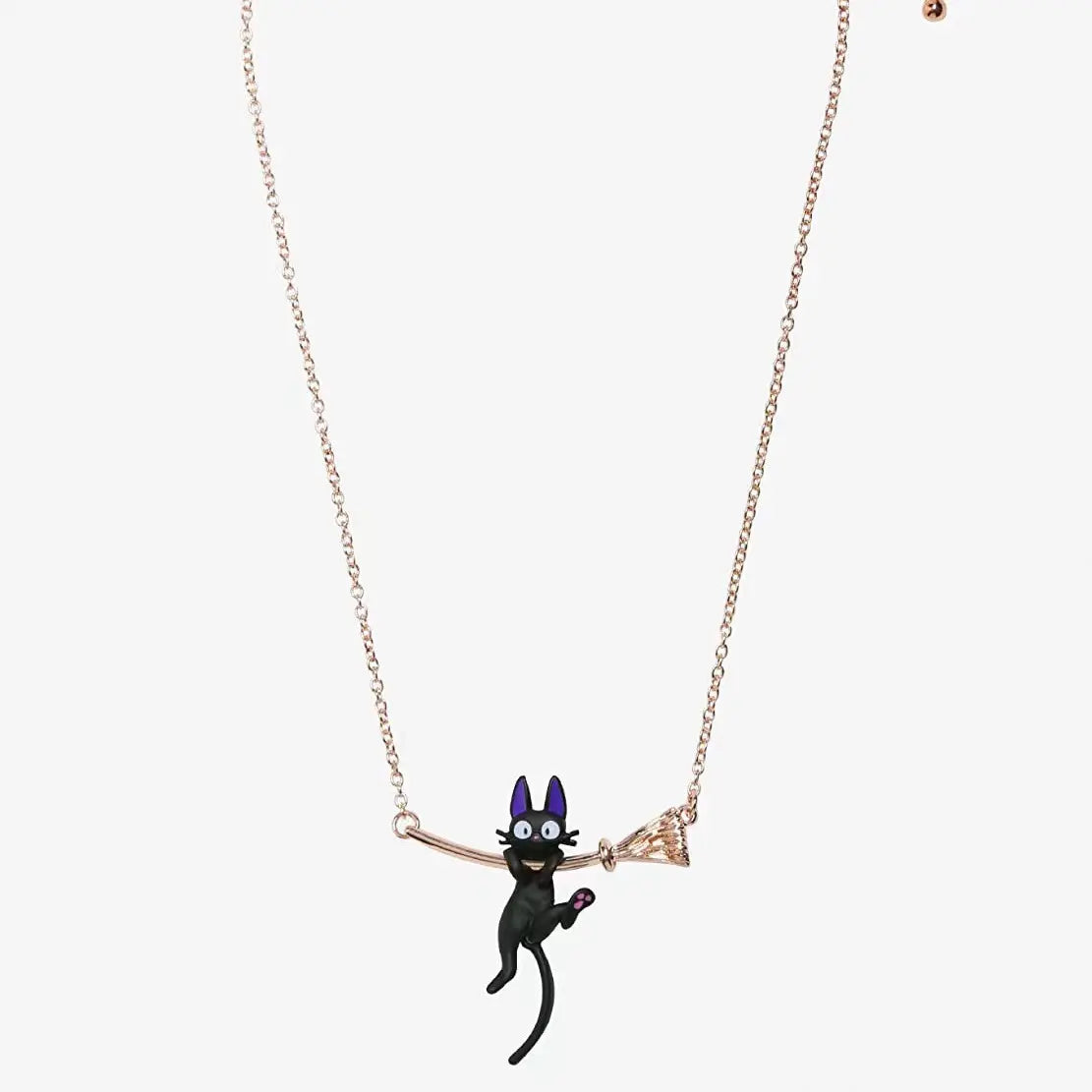 Close up of a necklace against a white background. The necklace includes a gold-colored witch's broomstick, with a black cat hanging from the handle. The inside of the cat's ears are purple.